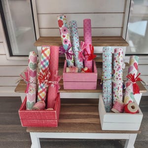 Puede incluir: Una exhibición de rollos de papel de regalo decorativos con varios diseños, incluyendo corazones y flores. Los rollos están dispuestos en cajas de madera rosas y blancas, con carretes de hilo y detalles decorativos.