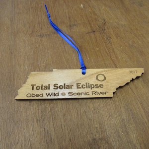 May include: A wooden ornament shaped like the state of Tennessee with the text "Total Solar Eclipse 2017 Obed Wild & Scenic River" engraved on it. The ornament has a blue ribbon attached to it.