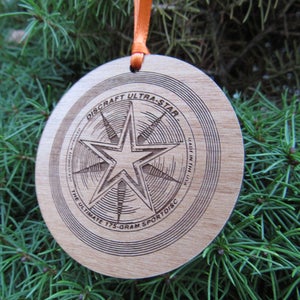 May include: A wooden disc ornament with a laser-engraved design featuring a five-pointed star and the text "DISCRAFT ULTRA-STAR. THE ULTIMATE 175-GRAM SPORTDISC. MADE IN THE USA."