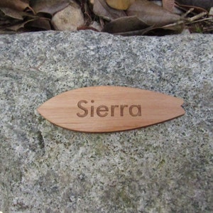 May include: A wooden surfboard-shaped ornament with the name "Sierra" engraved on it. The ornament is resting on a gray rock.