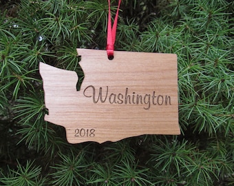 May include: Wooden Christmas ornament shaped like the state of Washington with the word "Washington" and the year "2018" engraved on it. The ornament is hanging from a red ribbon on a green pine tree.