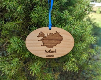 May include: A wooden ornament shaped like the island of Iceland with the year 2022 engraved below. The ornament is hanging from a blue ribbon against a green evergreen tree background.