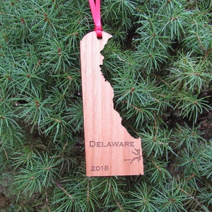 May include: A wooden ornament shaped like the state of Delaware with the word "DELAWARE" and the year "2018" engraved on it. The ornament is hanging from a red ribbon on a green evergreen tree.