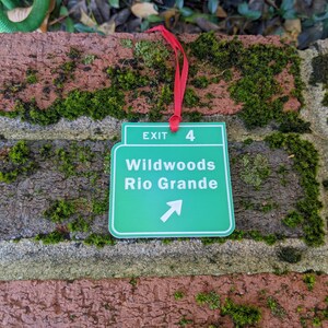 May include: Green exit sign with white text that reads "Exit 4 Wildwoods Rio Grande" with a white arrow pointing to the right. The sign is hanging from a red ribbon.