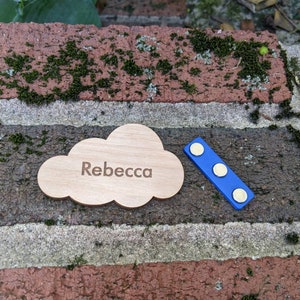 May include: A wooden cloud-shaped name tag with the name "Rebecca" engraved on it. The tag is attached to a blue magnetic strip with three gold-colored magnets.