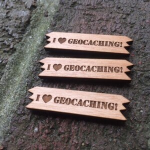 May include: Three wooden tags with the phrase "I heart geocaching!" engraved. Each tag has a heart symbol and a banner shape with pointed ends. The tags are light brown and rest on a textured, dark surface.