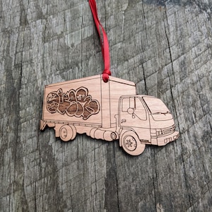 May include: Wooden Christmas ornament shaped like a delivery truck with graffiti on the side. The truck is brown and has a red ribbon attached to the top.