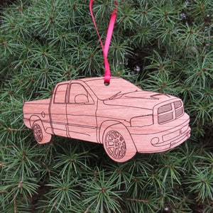 May include: A wooden Christmas ornament shaped like a Dodge Ram pickup truck. The ornament is painted in a light brown color and has a red ribbon attached to the top.