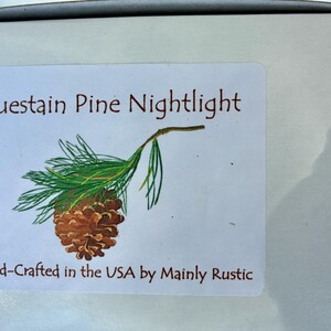 May include: A white label with the text "Bluestain Pine Nightlight" in brown lettering, accompanied by a detailed illustration of a pine cone and needles. The label also includes the text "Hand-Crafted in the USA by Mainly Rustic."