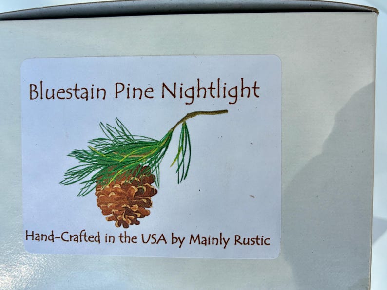 May include: A white label with the text "Bluestain Pine Nightlight" in brown lettering, accompanied by a detailed illustration of a pine cone and needles. The label also includes the text "Hand-Crafted in the USA by Mainly Rustic."