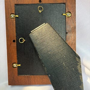 May include: A rectangular wooden picture frame shown from the back. It features a dark brown wood border, a black textured backing panel, and a hinged black easel stand. Gold-toned metal swivel clips secure the backing to the frame.