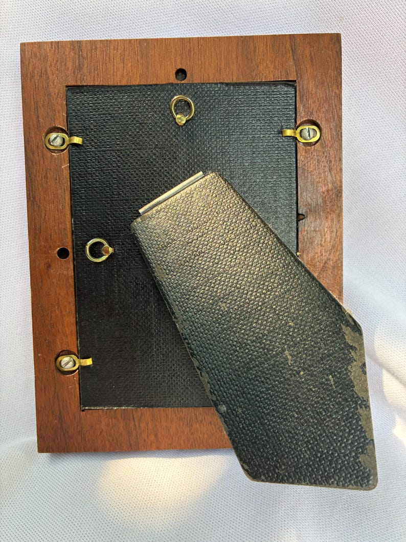 May include: A rectangular wooden picture frame shown from the back. It features a dark brown wood border, a black textured backing panel, and a hinged black easel stand. Gold-toned metal swivel clips secure the backing to the frame.
