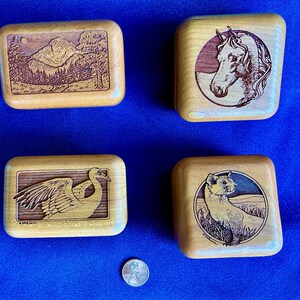 May include: Four small, rectangular wooden boxes with carved designs. One box features a mountain landscape, another a horse's head, a third a crane, and the last a weasel. A penny is included for size reference.