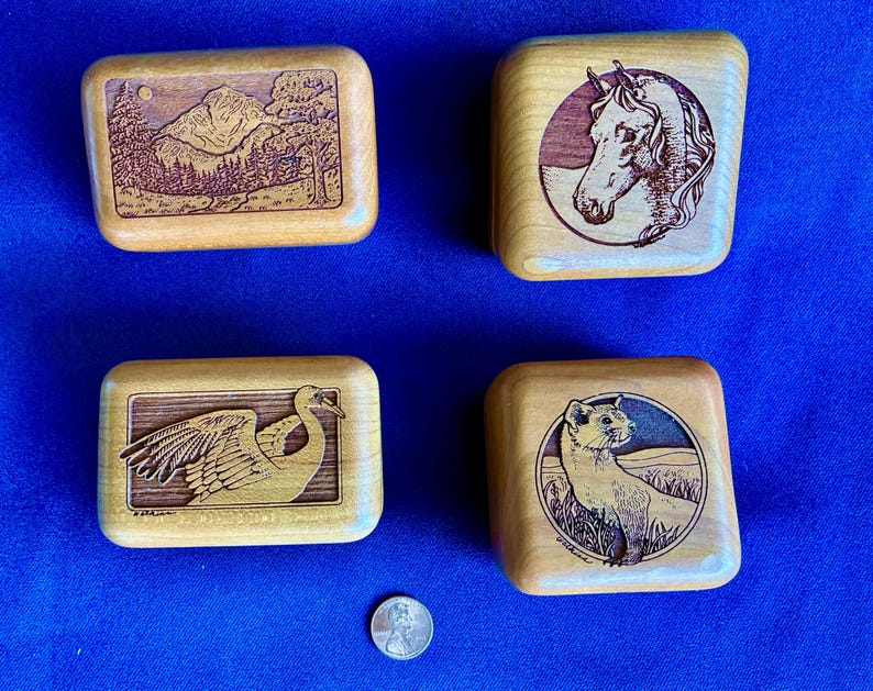 May include: Four small, rectangular wooden boxes with carved designs. One box features a mountain landscape, another a horse's head, a third a crane, and the last a weasel. A penny is included for size reference.