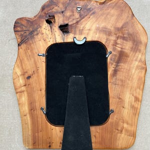 May include: A rectangular picture frame crafted from a natural live-edge wood slab with a polished finish. The frame features a black velvet backing, a central easel stand, four metal swivel clips, and a metal hanging hook at the top center.