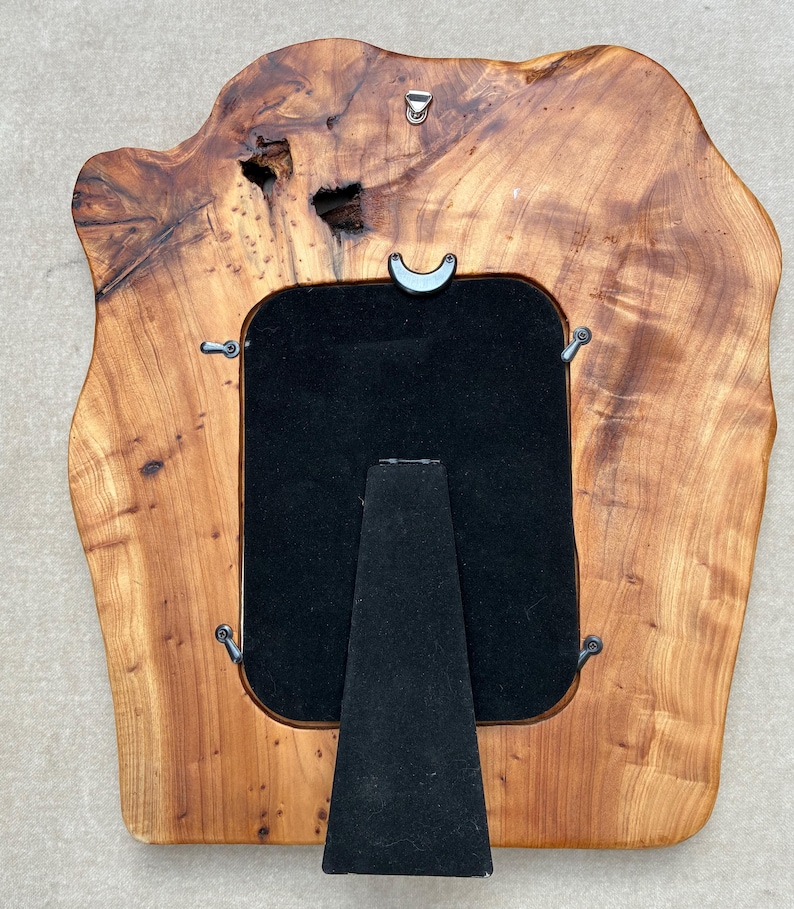May include: A rectangular picture frame crafted from a natural live-edge wood slab with a polished finish. The frame features a black velvet backing, a central easel stand, four metal swivel clips, and a metal hanging hook at the top center.