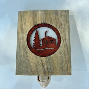 May include: A wooden night light with a rustic design. The light features a rectangular wooden frame with a circular cutout showcasing a cabin and tree silhouette in a reddish-brown hue. The night light has a standard plug-in base.