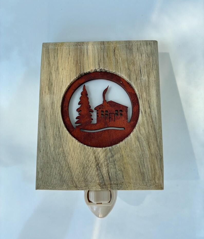 May include: A wooden night light with a rustic design. The light features a rectangular wooden frame with a circular cutout showcasing a cabin and tree silhouette in a reddish-brown hue. The night light has a standard plug-in base.