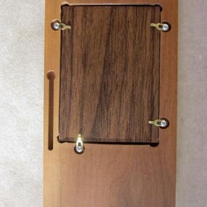 May include: A rectangular wooden frame backing with a light brown outer border and a darker brown wood grain center panel. The assembly features four small metal swivel clips, a single metal hanging hook, and two recessed keyhole slots for mounting.