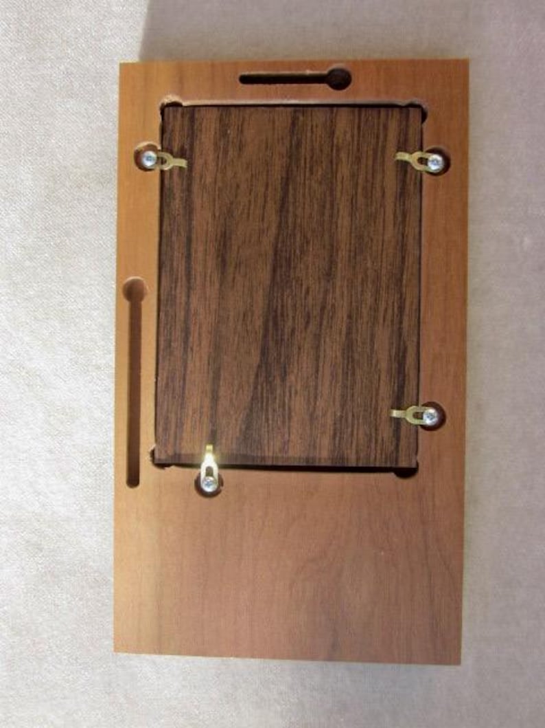 May include: A rectangular wooden frame backing with a light brown outer border and a darker brown wood grain center panel. The assembly features four small metal swivel clips, a single metal hanging hook, and two recessed keyhole slots for mounting.