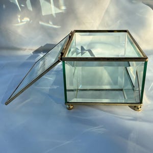 May include: A clear glass box with a hinged lid, featuring a gold-colored metal frame and small ball feet. The lid is partially open, revealing the interior. The box is designed to hold small items and is suitable for display.