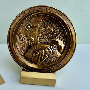 May include: A circular bronze-toned commemorative medallion featuring the text THE NEW MILLENNIUM and LOOKING TOWARD OUR FUTURE. The center depicts a relief of a globe, a stylized tree, a starburst, and the large embossed number 2000.
