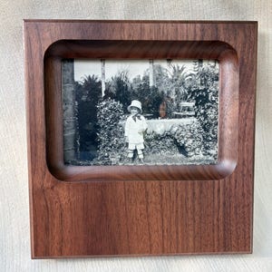 May include: A square, dark brown wooden picture frame with a smooth, recessed inner border. The frame holds a black and white photograph of a child wearing a hat and light-colored clothing, standing in a garden setting with trees and foliage.