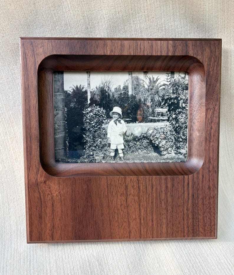 May include: A square, dark brown wooden picture frame with a smooth, recessed inner border. The frame holds a black and white photograph of a child wearing a hat and light-colored clothing, standing in a garden setting with trees and foliage.