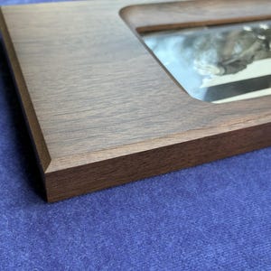 May include: A rectangular dark brown wooden frame with a smooth, matte finish and beveled edges. The frame features a recessed center opening that holds a reflective glass pane, showcasing a blurred, monochromatic image visible underneath.