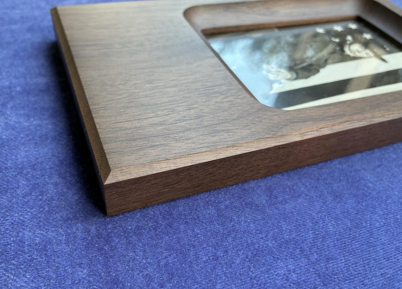 May include: A rectangular dark brown wooden frame with a smooth, matte finish and beveled edges. The frame features a recessed center opening that holds a reflective glass pane, showcasing a blurred, monochromatic image visible underneath.