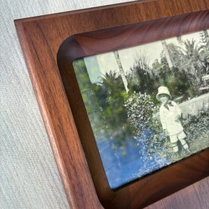 May include: A rectangular wooden picture frame with a smooth, dark brown finish and a recessed center. The frame holds a vintage-style photograph featuring a child wearing a hat and light-colored clothing, standing in a garden with palm trees.