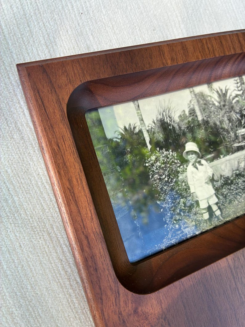 May include: A rectangular wooden picture frame with a smooth, dark brown finish and a recessed center. The frame holds a vintage-style photograph featuring a child wearing a hat and light-colored clothing, standing in a garden with palm trees.
