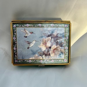 May include: A rectangular brass trinket box featuring a hinged lid with an enamel inlay. The lid displays two hummingbirds in flight near large, light-colored hibiscus flowers against a blue background, framed by a green floral border.