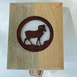 May include: A wooden night light featuring a circular, rusty-brown ram silhouette. The ram is set against a white background within a wooden frame, with a two-tone finish. The night light has a standard plug.