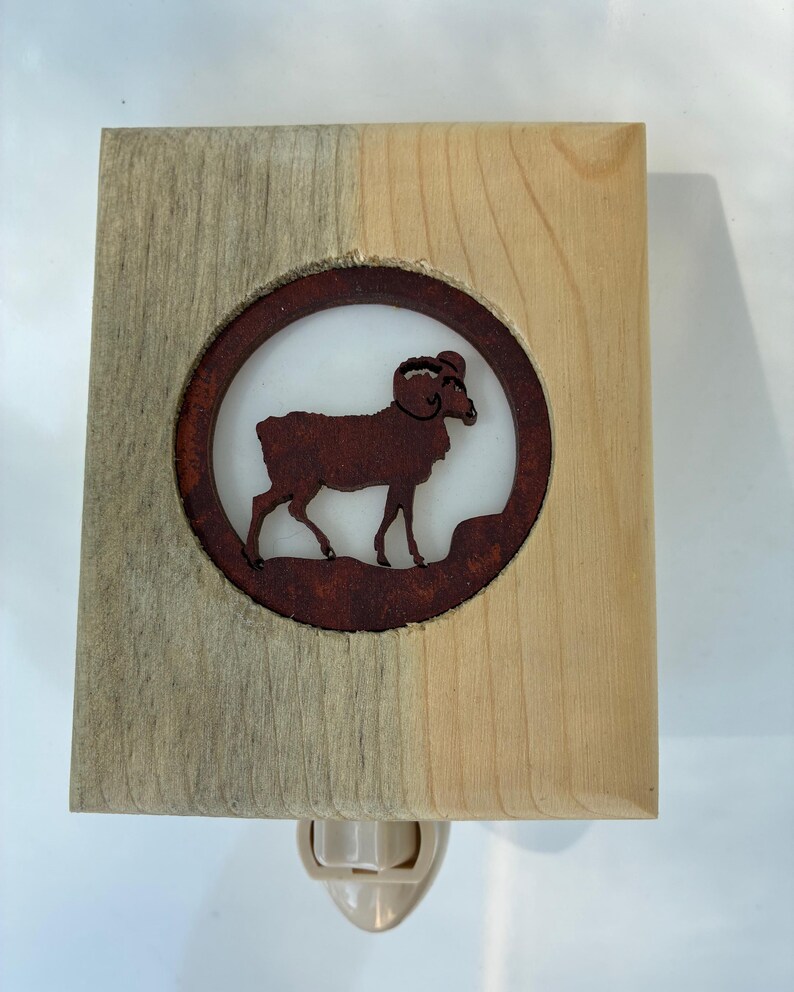 May include: A wooden night light featuring a circular, rusty-brown ram silhouette. The ram is set against a white background within a wooden frame, with a two-tone finish. The night light has a standard plug.