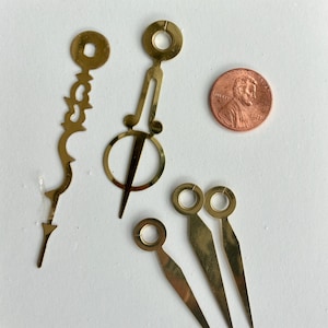 May include: A selection of brass clock hands and components, including decorative hour and minute hands, and a penny for scale. The hands have a gold finish and are arranged on a white surface. The clock parts are likely for a DIY or repair project.
