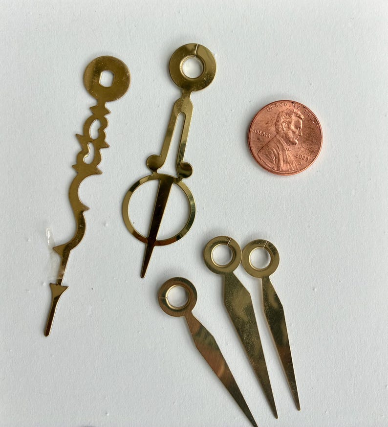 May include: A selection of brass clock hands and components, including decorative hour and minute hands, and a penny for scale. The hands have a gold finish and are arranged on a white surface. The clock parts are likely for a DIY or repair project.
