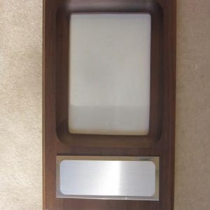 May include: A rectangular wooden plaque featuring a dark brown finish with a recessed vertical white center panel and a horizontal silver-toned metal plate positioned below the white area, designed for mounting or display purposes.