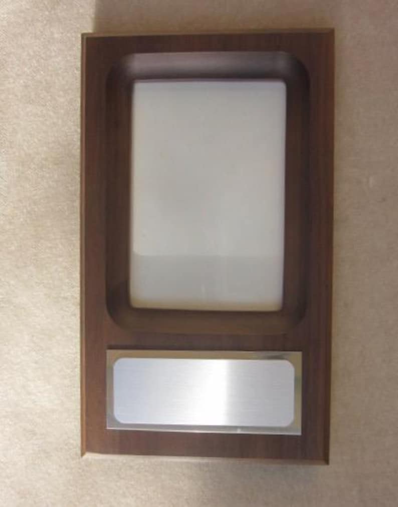 May include: A rectangular wooden plaque featuring a dark brown finish with a recessed vertical white center panel and a horizontal silver-toned metal plate positioned below the white area, designed for mounting or display purposes.
