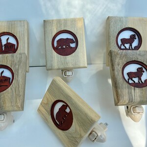 May include: Wooden night lights with a natural wood grain finish. Each light features a circular cutout with a brown silhouette of a bear, ram, or cabin scene. The lights have a white plug-in base and are designed for use in a wall outlet.
