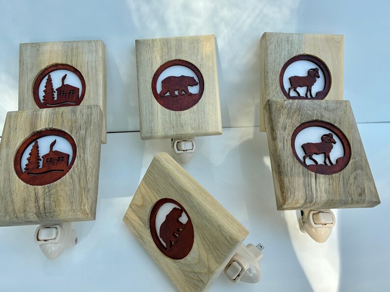 May include: Wooden night lights with a natural wood grain finish. Each light features a circular cutout with a brown silhouette of a bear, ram, or cabin scene. The lights have a white plug-in base and are designed for use in a wall outlet.