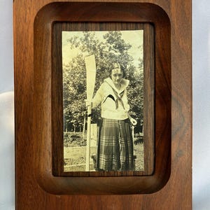 May include: A rectangular wooden picture frame with a dark brown finish and a recessed center. Inside the frame is a black and white photograph of a person wearing a sailor-style collared shirt and a plaid skirt, holding a long wooden oar upright.