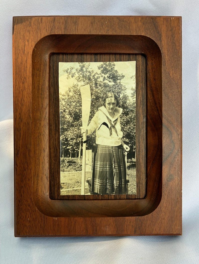 May include: A rectangular wooden picture frame with a dark brown finish and a recessed center. Inside the frame is a black and white photograph of a person wearing a sailor-style collared shirt and a plaid skirt, holding a long wooden oar upright.