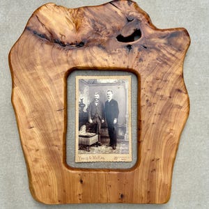May include: A live edge wooden picture frame with a natural grain pattern and a dark knot detail at the top. The frame holds a vintage black and white photograph of two individuals standing in a room, with the text Young & McKay, Marshall, Mich.