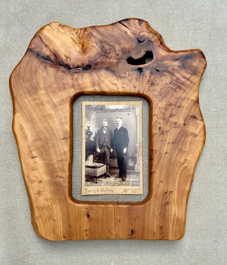 May include: A live edge wooden picture frame with a natural grain pattern and a dark knot detail at the top. The frame holds a vintage black and white photograph of two individuals standing in a room, with the text Young & McKay, Marshall, Mich.