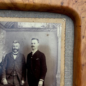 May include: A vintage black and white photograph of two men in suits, one with a prominent mustache, set within a polished wooden frame featuring a distinct burl wood grain pattern and a textured gray border surrounding the scalloped-edge photo.