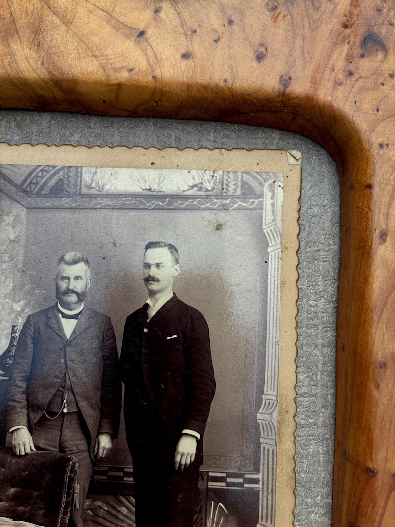 May include: A vintage black and white photograph of two men in suits, one with a prominent mustache, set within a polished wooden frame featuring a distinct burl wood grain pattern and a textured gray border surrounding the scalloped-edge photo.