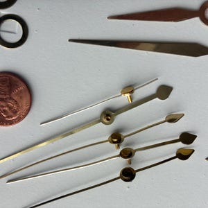 May include: Close-up of various clock hands in gold, silver, and copper tones. A penny is included for size reference. The hands have different shapes, including pointed, leaf, and circular designs. A few small gold rings are also visible.