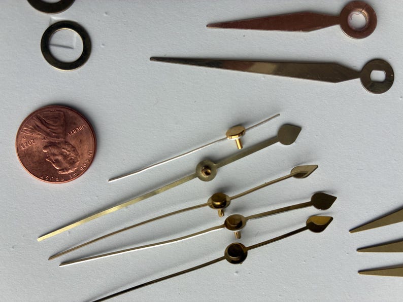 May include: Close-up of various clock hands in gold, silver, and copper tones. A penny is included for size reference. The hands have different shapes, including pointed, leaf, and circular designs. A few small gold rings are also visible.