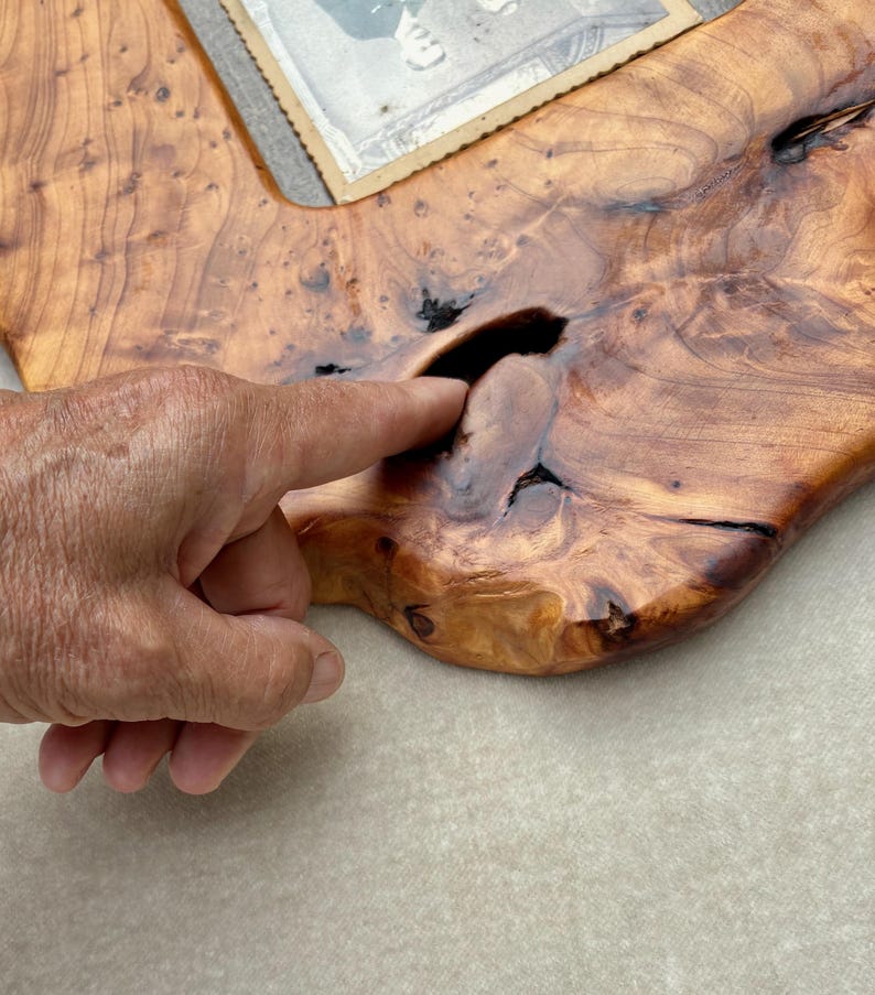 May include: A polished wooden picture frame crafted from a natural slab with a prominent live edge and visible wood grain. The frame features a dark, irregular knot hole in the center of the wood surface and holds a vintage black and white photograph.
